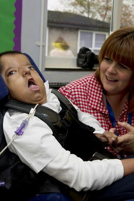 A young boy in a supportive chair
                  holds one of the finger's of his teacher's hand