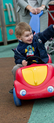 A boy drives a toy car around a playground,
pushed by an adult