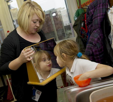 A young girl looks at herself
                  in a mirror held by a member of staff