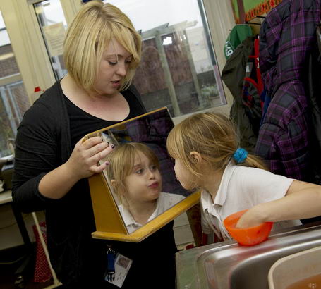 A young girl looks at herself
                  in a mirror held by a member of staff