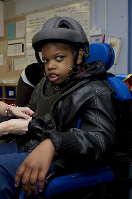 A young black boy with protective
                  headgear in a wheelchair is helped by a teacher