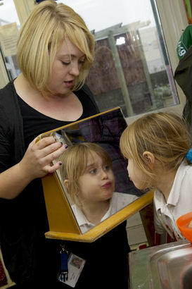 A young girl looks at herself
                  in a mirror held by a member of staff