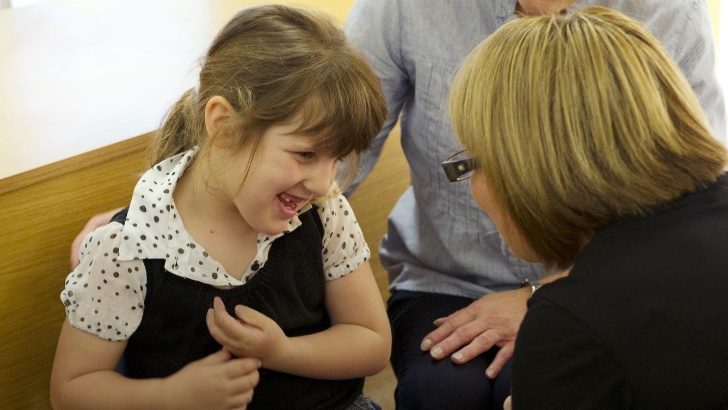 A seated girl smiles as a teacher
                  talks to her