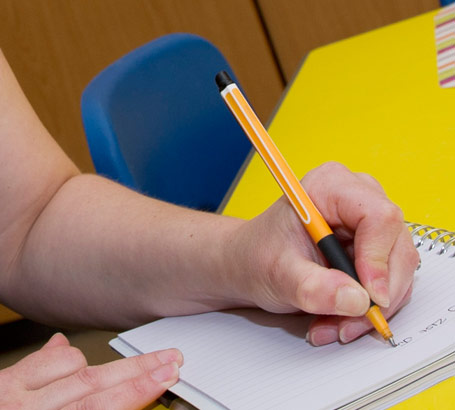 a teacher wearing glasses sits
at a table taking notes on one of her pupils