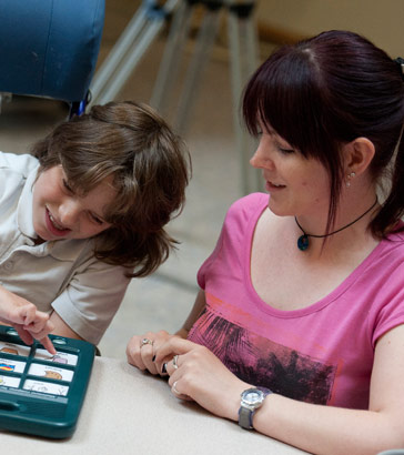 A teacher watches a boy select an
                  image on a communication device