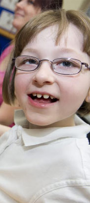 young
girl in white top smiles at camera, sitting next to assistive device