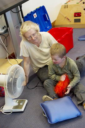 A teacher and a young boy seated
on the floor with a cushion and a fan
