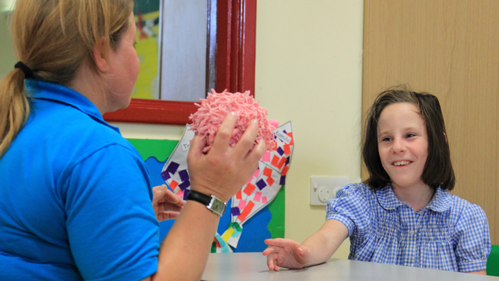 A girl sitting opposite her
                  teacher smiles as the teacher holds a pink toy