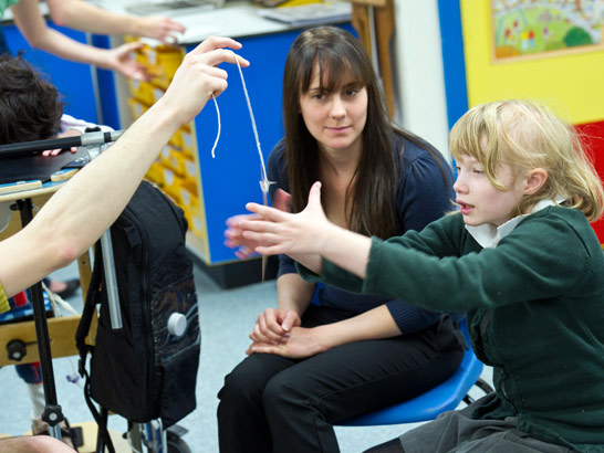 a young girl spins a pendulum
whilst her teacher and classmates look on