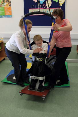 Two carers help a boy into a standing
                  support