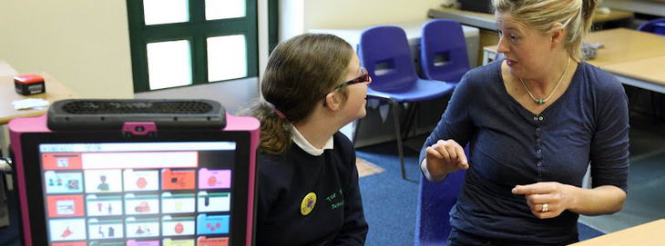 A teacher sits next to a girl with a communication
                  device