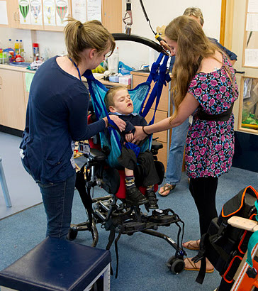 A boy being lifted out of a wheelchair
                  with a hoist