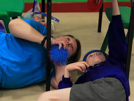 Carer sits under a table with a disabled
                  boy.